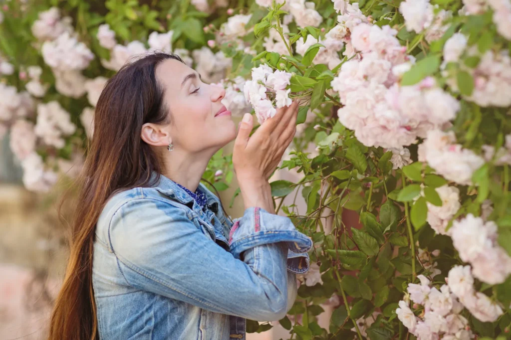 אישה מחייכת ומריחה פרחים ורודים, מוקפת בטבע יפהפה וריחני.